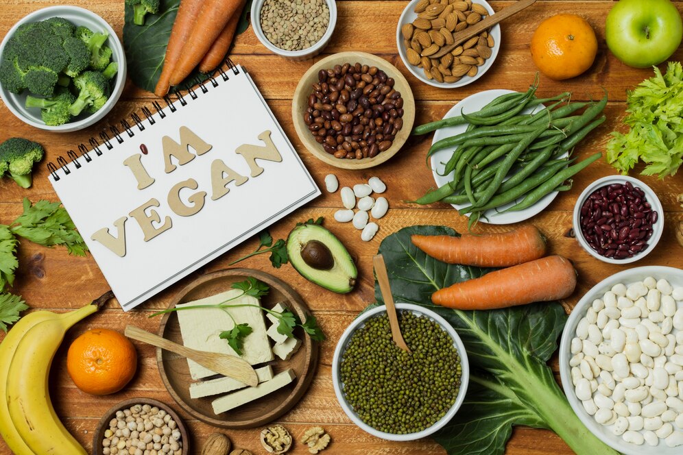 A flat lay of various vegan foods, including vegetables, fruits, legumes, and plant-based ingredients, with the words 'I'M VEGAN' spelled out in letter blocks on a notepad.