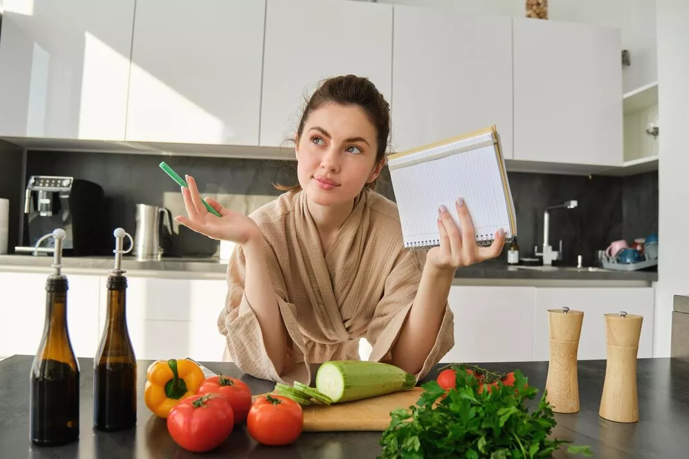 A woman in a kitchen holding a notepad and pen, looking thoughtfully at the ingredients on the table, including tomatoes, cucumbers, and fresh herbs, while considering what to cook.