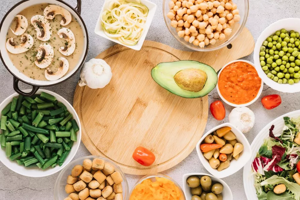 A variety of top plant-based anti-inflammatory foods, including avocado, chickpeas, green beans, peas, mushrooms, garlic, olives, fresh salad, and vegetable-based sauces, arranged around a wooden cutting board
