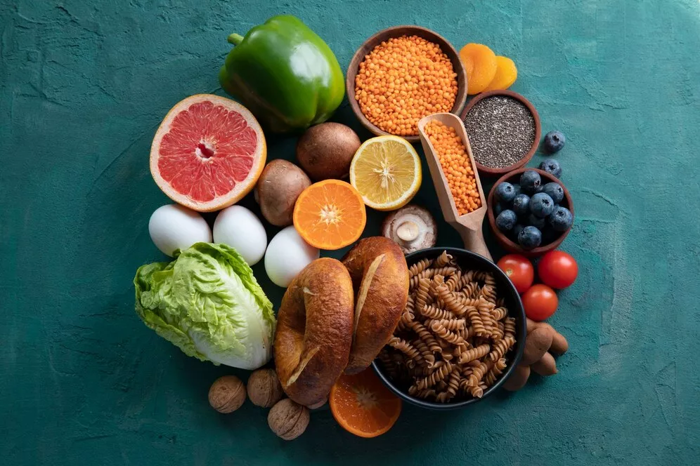 A selection of nutritious plant-based foods, including lentils, chia seeds, blueberries, tomatoes, citrus fruits, lettuce, whole grain pasta, and bread, arranged on a textured green background.