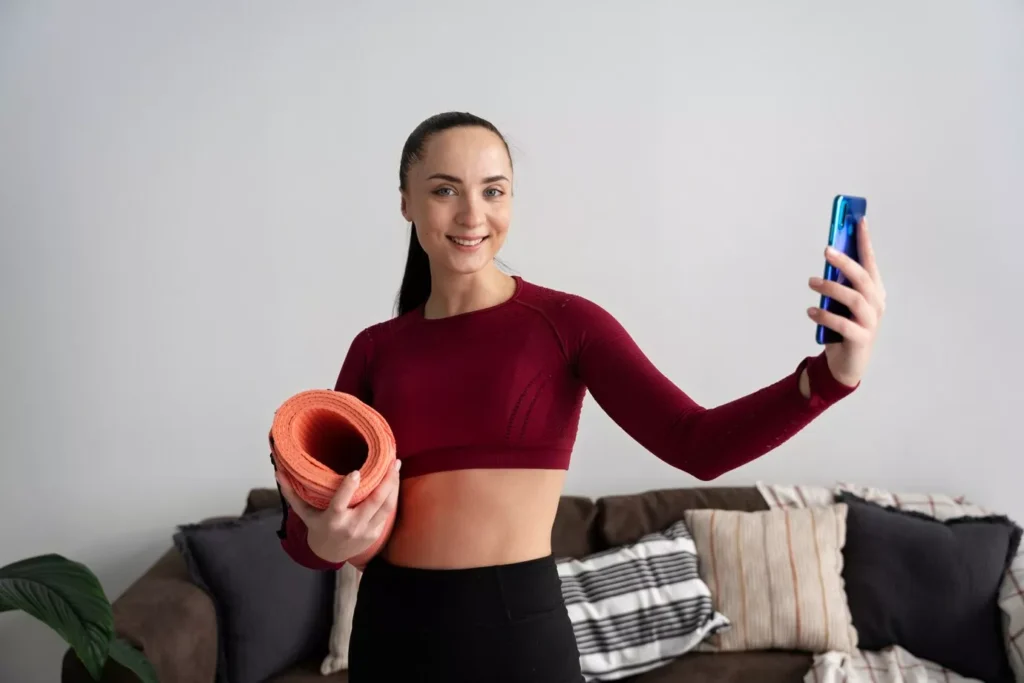 Smiling woman in workout attire holding a yoga mat and taking a selfie, promoting exercise for better digestion